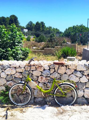 Side photo of a green Graziella leaning against a dry stone wall. Behind it is Mediterranean vegetation, the sky is blue