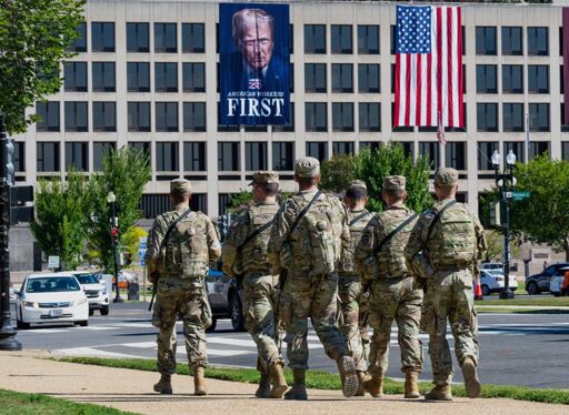 A group of U.S. soldiers in camouflage uniforms walk in formation along a city street. In the background, a government building displays a massive banner of Donald Trump with the slogan “American Workers First” beside a large American flag, evoking a scene of military presence and nationalism.
