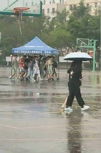 Two people walk under a umbrella while a whole herd of people scuffle along under one of those half tent things they have a conventions 
